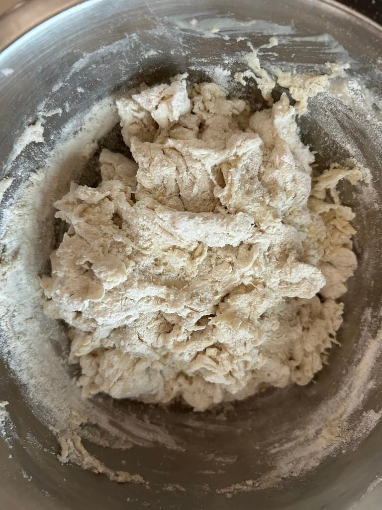 Flour and yeast mixture coming together into a rough, sticky bread dough in a stainless steel bowl.